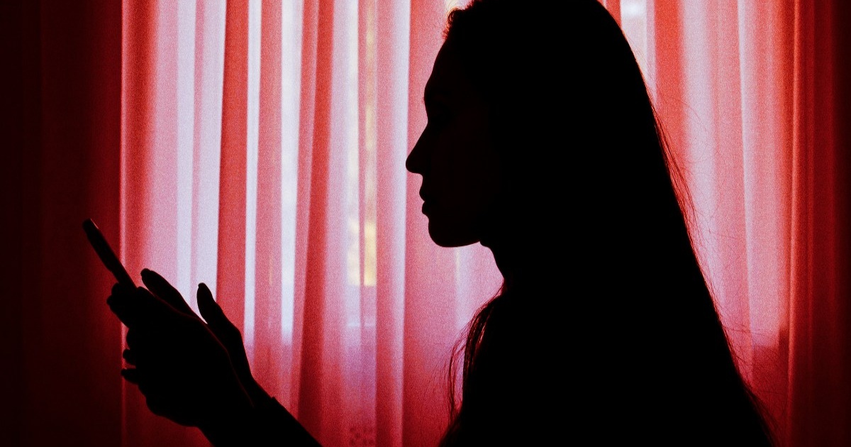 Siluet wanita duduk dalam gelap sambil memegang ponsel yang menyala, menggambarkan kesendirian pengguna yang berinteraksi dengan chatbot AI