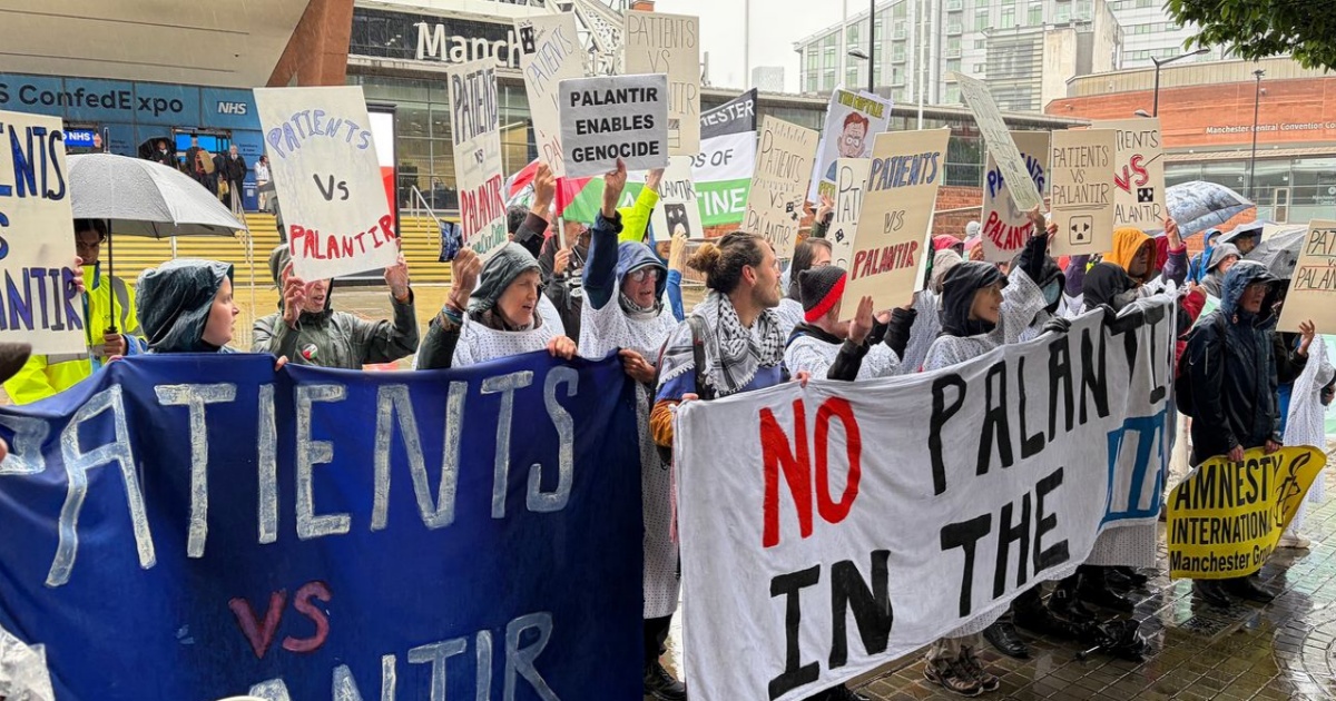 Pengunjuk rasa membawa poster bertuliskan Patients vs Palantir di depan NHS ConfedExpo Manchester