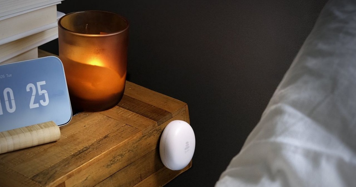 Perangkat Halo berbentuk puck putih kecil dengan logo ScreenZen, ditempatkan di atas meja nakas kayu di samping tempat tidur
